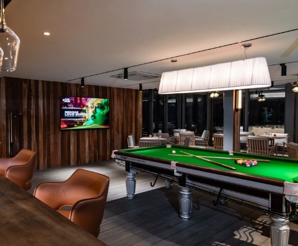 A stylish bar featuring a green pool table, modern bar seating, and an illuminated light fixture, with a TV displaying vibrant content.