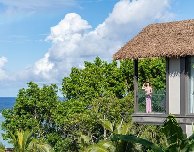A woman in a pink dress stands on a balcony overlooking the ocean, surrounded by lush greenery and a clear blue sky.