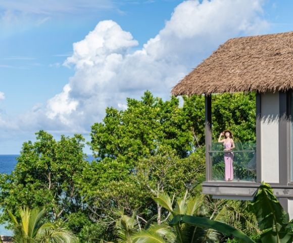 A woman in a pink dress stands on a balcony overlooking the ocean, surrounded by lush greenery and a clear blue sky.