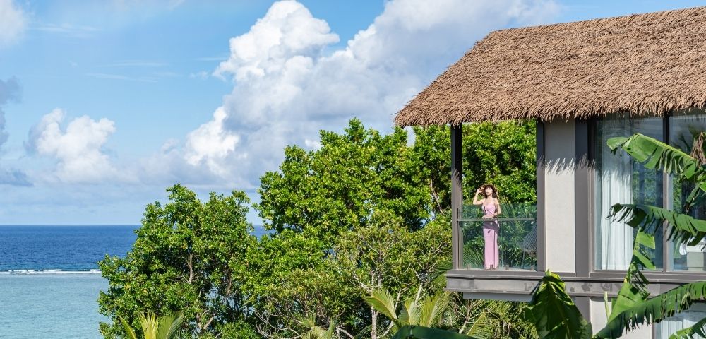 A woman in a pink dress stands on a balcony overlooking the ocean, surrounded by lush greenery and a clear blue sky.