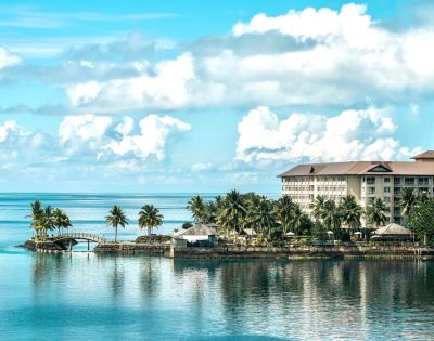 A serene waterfront resort with palm trees and a wooden bridge, set against a bright blue sky and calm ocean waters.