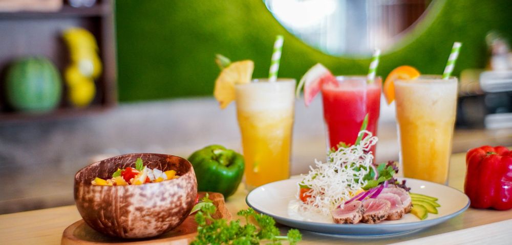 Colorful tropical drinks and fresh dishes served in wooden bowls, showcasing vibrant ingredients in a bright restaurant setting.