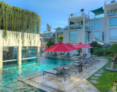 Lush pool area featuring modern architecture, loungers under red umbrellas, and tropical greenery in bright sunlight.