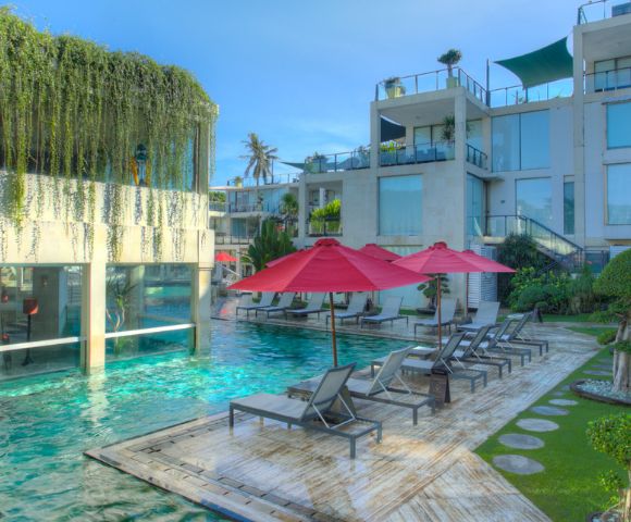 Lush pool area featuring modern architecture, loungers under red umbrellas, and tropical greenery in bright sunlight.