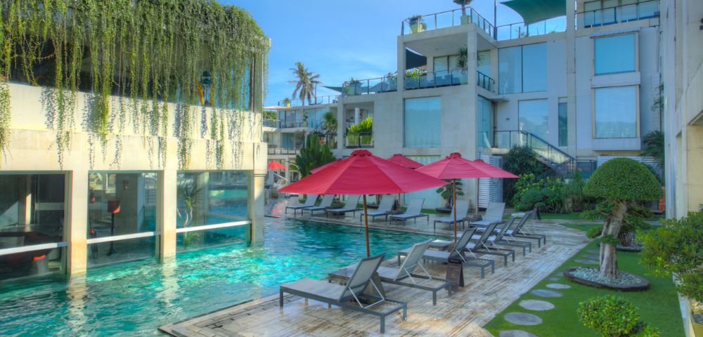 Lush pool area featuring modern architecture, loungers under red umbrellas, and tropical greenery in bright sunlight.