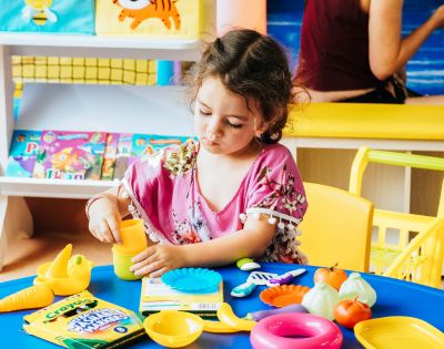 A child plays with colorful toy food and utensils on a blue table, surrounded by educational books in a bright, cheerful environment.
