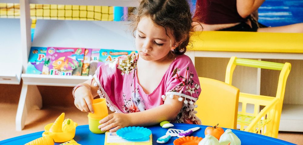 A child plays with colorful toy food and utensils on a blue table, surrounded by educational books in a bright, cheerful environment.