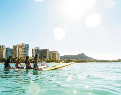 Group of surfers sitting on boards in turquoise water, with Honolulu skyscrapers and Diamond Head mountain in the background.