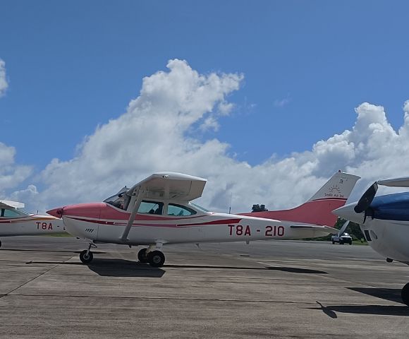 A Cessna aircraft with a red and white design, parked on a tarmac under a blue sky with fluffy clouds.
