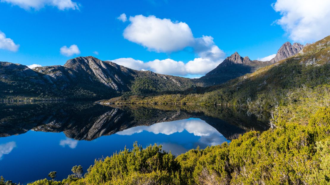 Serene landscape featuring a calm lake reflecting mountains and blue skies, surrounded by lush greenery and scattered clouds.