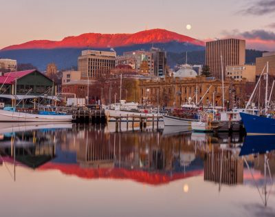 Sunset casts a warm glow on Mount Wellington, reflecting in the tranquil harbor with boats and city buildings in the foreground.