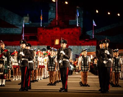 Uniformed military band members perform in formation outdoors at night, with pipers and dancers in traditional attire and a historic building in the background.