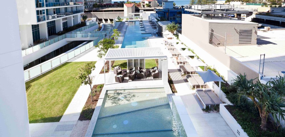 A captivating image displays a tranquil pool scene, where a pristine blue pool, framed by a white railing, is nestled among lush greenery and modern white buildings. The image captures the perfect blend of serenity and urban sophistication, inviting relaxation in every detail.