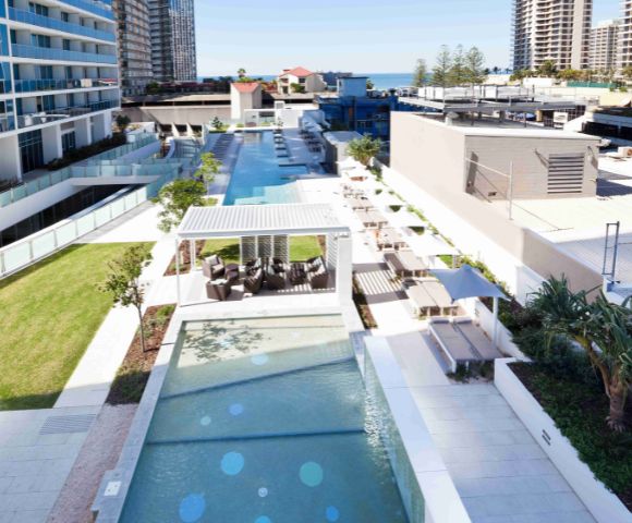 A captivating image displays a tranquil pool scene, where a pristine blue pool, framed by a white railing, is nestled among lush greenery and modern white buildings. The image captures the perfect blend of serenity and urban sophistication, inviting relaxation in every detail.