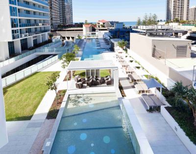 A captivating image displays a tranquil pool scene, where a pristine blue pool, framed by a white railing, is nestled among lush greenery and modern white buildings. The image captures the perfect blend of serenity and urban sophistication, inviting relaxation in every detail.