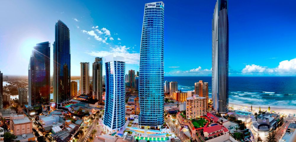 Image displays a harmonious blend of modern and traditional cityscape. The images focal point is a towering blue skyscraper, while the sea and sky provide a stunning backdrop. The red-roofed and diverse architectural details create a lively urban scene, captured in a serene and visually appealing composition.