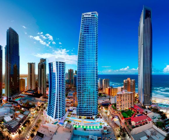 Image displays a harmonious blend of modern and traditional cityscape. The images focal point is a towering blue skyscraper, while the sea and sky provide a stunning backdrop. The red-roofed and diverse architectural details create a lively urban scene, captured in a serene and visually appealing composition.