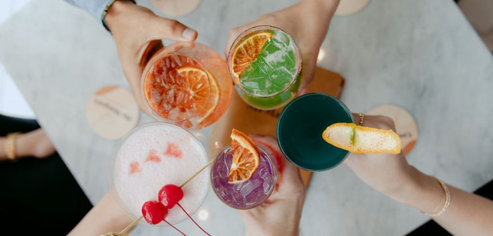 A captivating image displays a vivacious scene where four hands, holding drinks of diverse colors and designs, form a harmonious circle around a pristine white table. The array of clear and colored beverages, garnished with personal flair, fosters a sense of camaraderie and celebration.