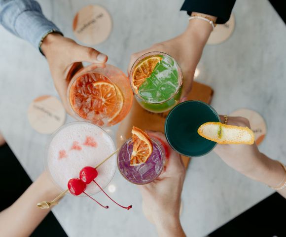 A captivating image displays a vivacious scene where four hands, holding drinks of diverse colors and designs, form a harmonious circle around a pristine white table. The array of clear and colored beverages, garnished with personal flair, fosters a sense of camaraderie and celebration.