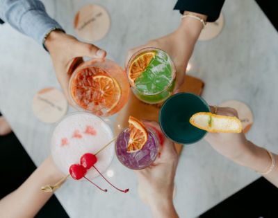 A captivating image displays a vivacious scene where four hands, holding drinks of diverse colors and designs, form a harmonious circle around a pristine white table. The array of clear and colored beverages, garnished with personal flair, fosters a sense of camaraderie and celebration.