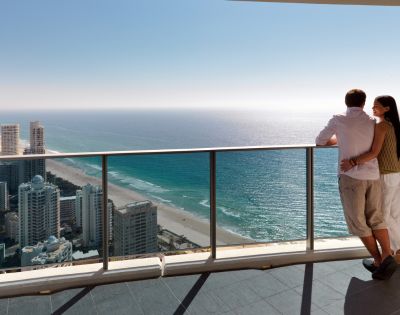 A couple stands on a balcony overlooking a vibrant blue ocean and sunny beach with a skyline of tall buildings in the background.