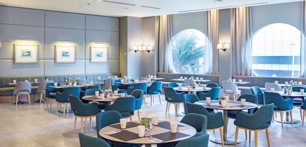 A captivating image displays a modern restaurant, where large windows and soothing blue furniture create a design masterpiece. The images elegance shines through the well-lit space, framed pictures, and a harmonious color palette that captivates the eye.