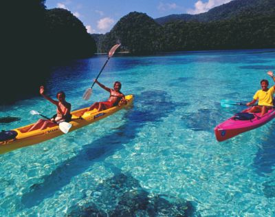 Two people paddle kayaks in clear turquoise waters, surrounded by lush green hills under a bright blue sky, enjoying a sunny day.