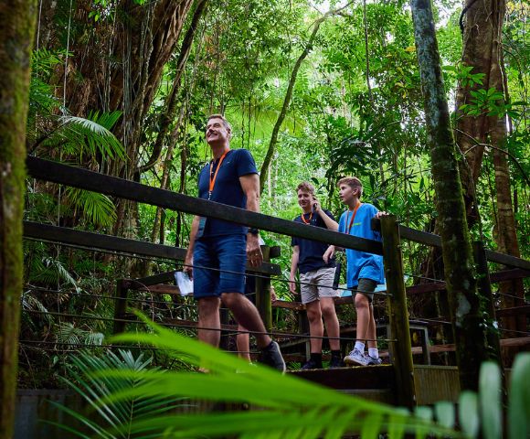 A group of people is walking on a bridge in a wooded area. The scene depicts individuals dressed for outdoor activities, such as hiking, with some wearing shorts. Surrounding them are trees and lush greenery typical of a forest or jungle environment, creating a tranquil vacation atmosphere.