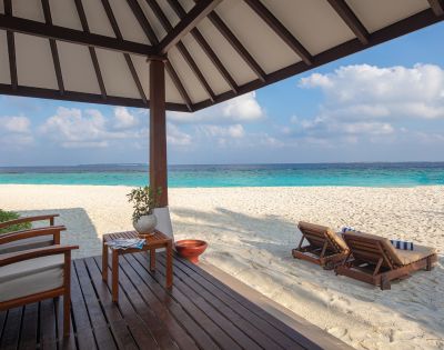 Cozy beachside bungalow with wooden chairs overlooks a serene, turquoise ocean. Two wooden loungers rest on white sand under a partly cloudy sky.