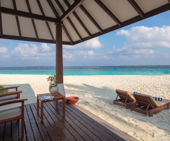 Cozy beachside bungalow with wooden chairs overlooks a serene, turquoise ocean. Two wooden loungers rest on white sand under a partly cloudy sky.