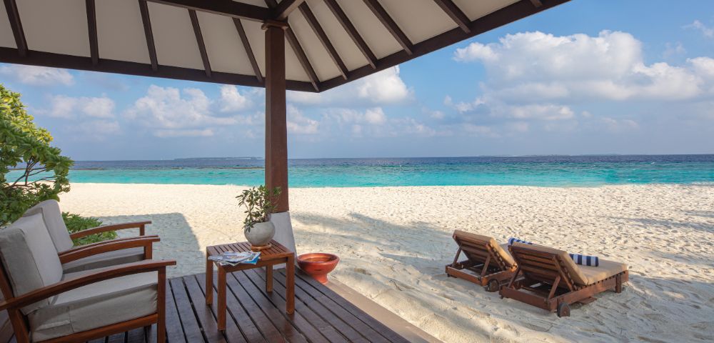 Cozy beachside bungalow with wooden chairs overlooks a serene, turquoise ocean. Two wooden loungers rest on white sand under a partly cloudy sky.