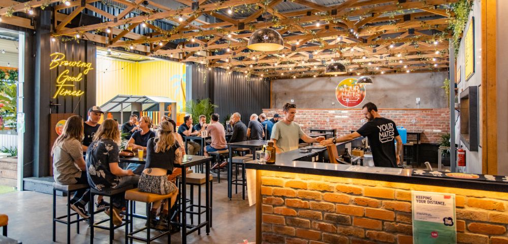 A bustling brewery with patrons enjoying drinks at high tables under a wooden ceiling, adorned with string lights and greenery.