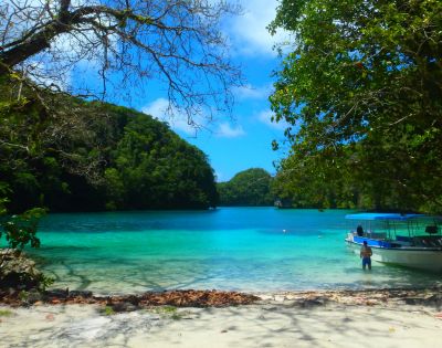 A secluded beach with turquoise water, lush green hills, and a small boat, while a person stands in the shallow water under a sunny sky.