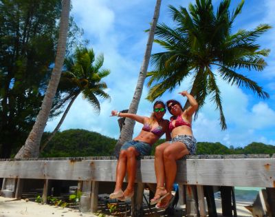 Two women in bikinis sit on a wooden platform surrounded by palm trees, smiling and posing against a tropical backdrop.