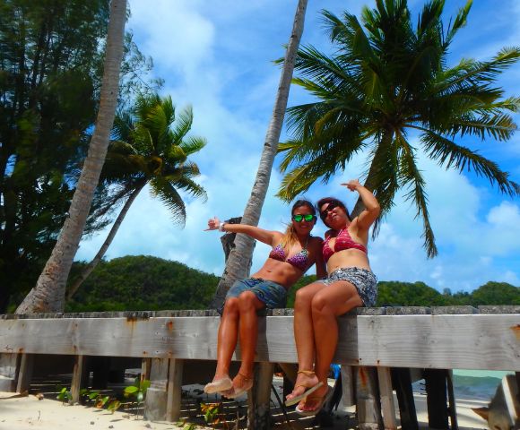 Two women in bikinis sit on a wooden platform surrounded by palm trees, smiling and posing against a tropical backdrop.