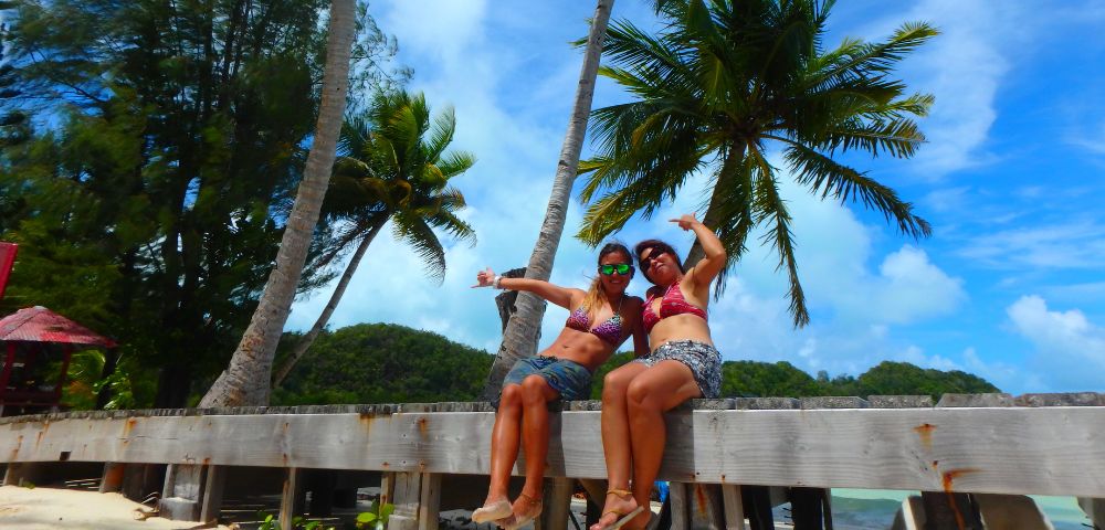 Two women in bikinis sit on a wooden platform surrounded by palm trees, smiling and posing against a tropical backdrop.
