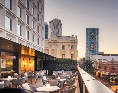 Modern rooftop terrace with cozy seating and glowing candle lights, overlooking a cityscape of historic and modern buildings. Soft evening light.