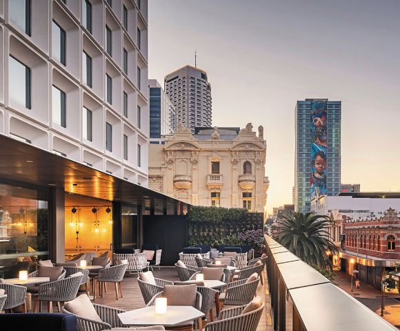 Modern rooftop terrace with cozy seating and glowing candle lights, overlooking a cityscape of historic and modern buildings. Soft evening light.