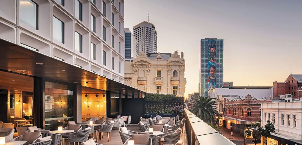 Modern rooftop terrace with cozy seating and glowing candle lights, overlooking a cityscape of historic and modern buildings. Soft evening light.
