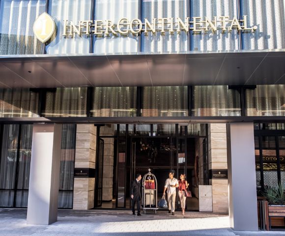 The exterior of an Intercontinental hotel showcases a sleek design with large glass doors and an inviting entryway. Guests are seen arriving, accompanied by a concierge holding a luggage cart.