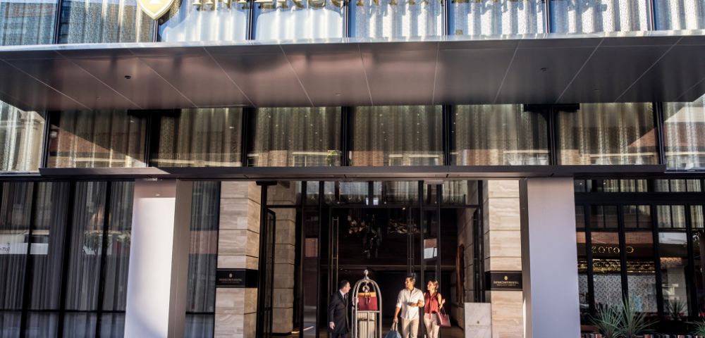 The exterior of an Intercontinental hotel showcases a sleek design with large glass doors and an inviting entryway. Guests are seen arriving, accompanied by a concierge holding a luggage cart.
