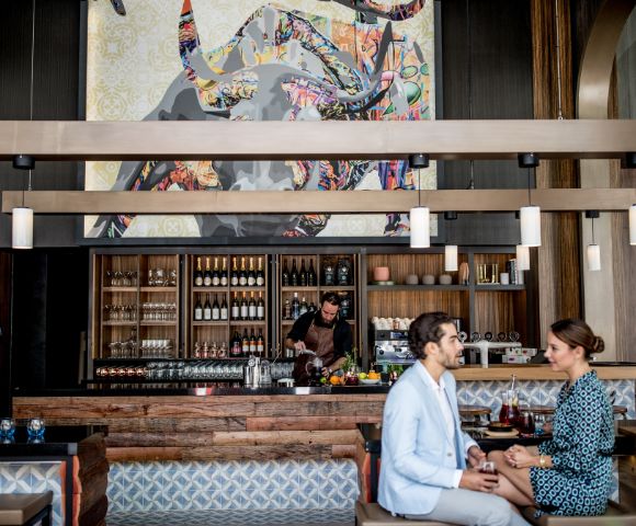 A couple is seated at a stylish bar, holding drinks while engaging in conversation. In the background, a bartender is preparing beverages against a backdrop of decorative shelves filled with bottles.