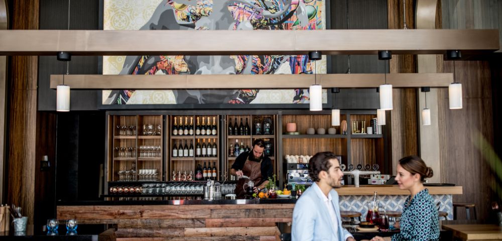 A couple is seated at a stylish bar, holding drinks while engaging in conversation. In the background, a bartender is preparing beverages against a backdrop of decorative shelves filled with bottles.