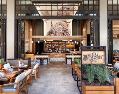 A modern restaurant interior featuring a stylish bar area and wooden tables with blue accents. An artistic mural hangs above the bar, enhancing the inviting atmosphere of the space.