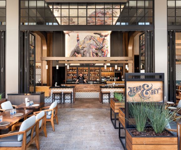 A modern restaurant interior featuring a stylish bar area and wooden tables with blue accents. An artistic mural hangs above the bar, enhancing the inviting atmosphere of the space.