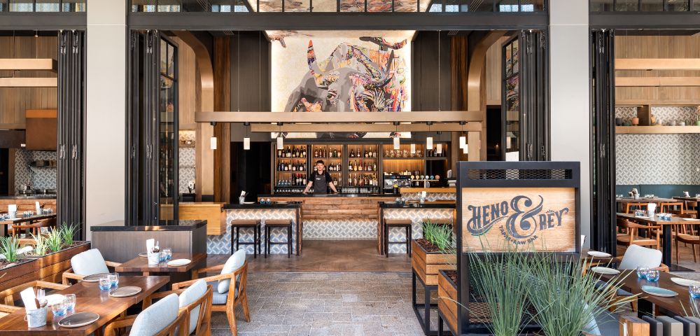 A modern restaurant interior featuring a stylish bar area and wooden tables with blue accents. An artistic mural hangs above the bar, enhancing the inviting atmosphere of the space.
