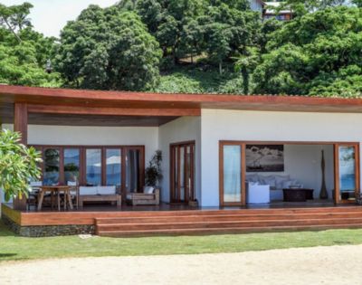 Modern beachfront house with wooden accents, large windows, and a spacious porch, surrounded by lush greenery.