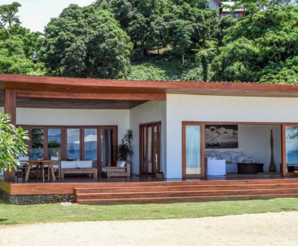 Modern beachfront house with wooden accents, large windows, and a spacious porch, surrounded by lush greenery.