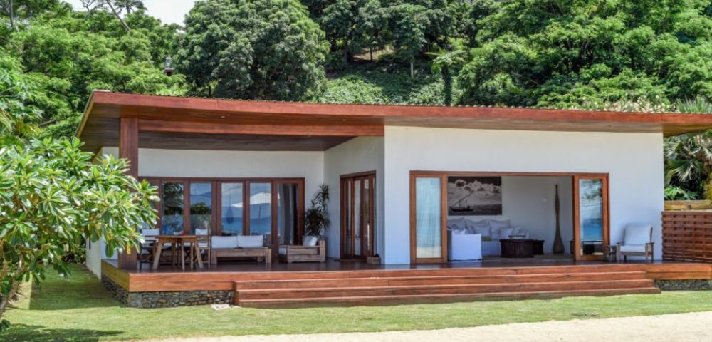 Modern beachfront house with wooden accents, large windows, and a spacious porch, surrounded by lush greenery.