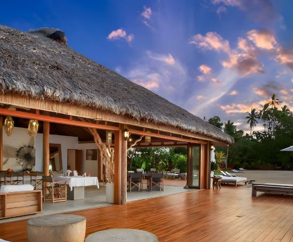 Tropical villa with a thatched roof, wooden deck, dining area, and beach view under a colorful sunset sky, surrounded by palm trees.